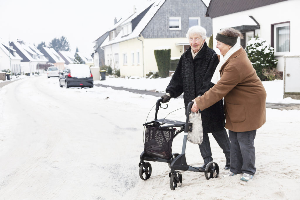 senior woman crossing road with the help a mature woman, using walker, having difficulties cause road is covered with snow