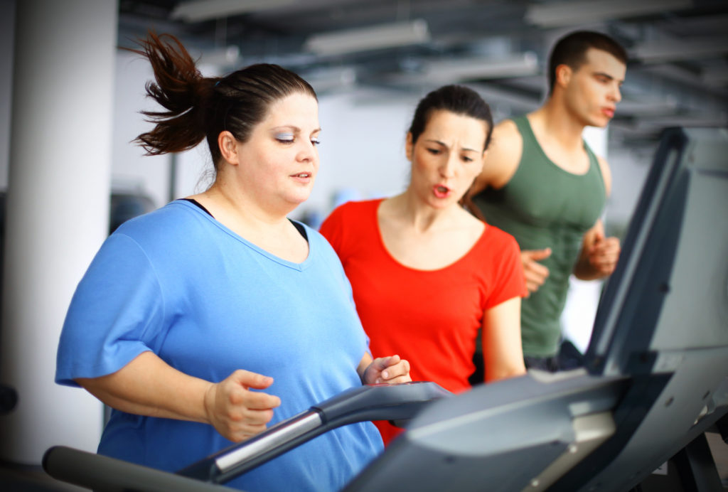 Overweight woman doing cardio workout on a treadmill in gym with her coach assisting and supporting her