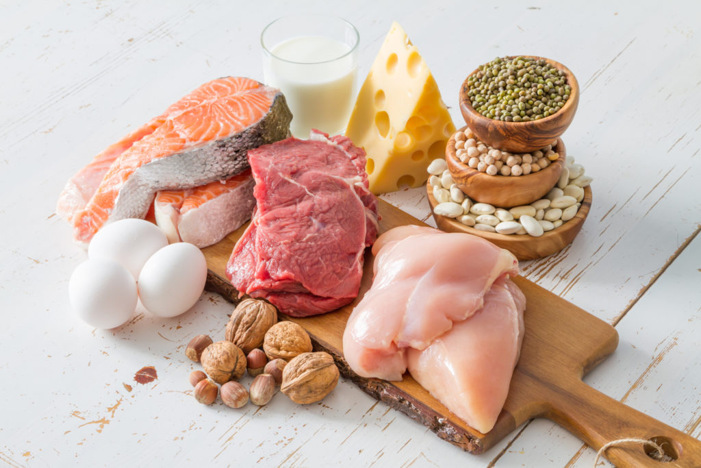 shot of chicken, beef and salmon on cutting board, surrounded by walnuts, eggs, cheese, glass of milk and other protein sources