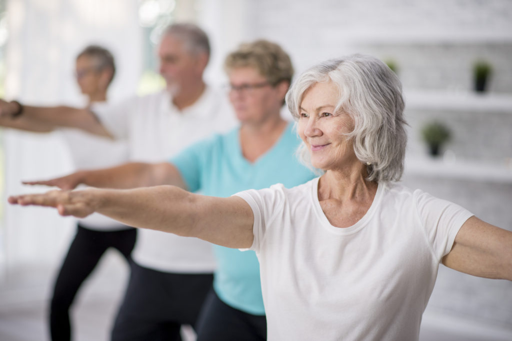 A multi-ethnic group of adult men and women are indoors in a fitness studio. They are wearing casual clothing while at a yoga class. A senior Caucasian woman is smiling while stretching out her arms.