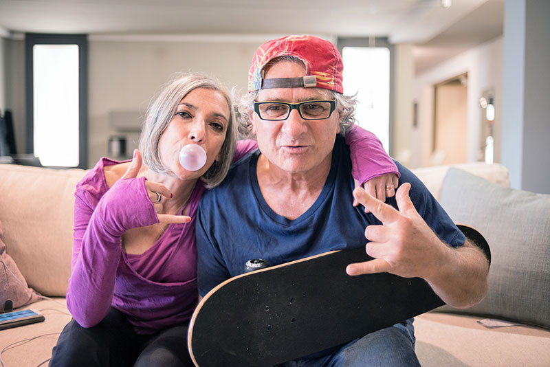 elder couple with youthful attitude. Woman is blowing a bubble gum bubble and man is holding a skateboard and wearing a backwards baseball cap