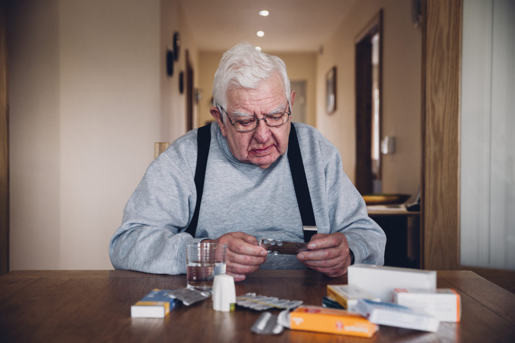 Elderly man sitting at home at a table with his head resting on his hand. Looking troubled he holds a packet of pills with other medication around him