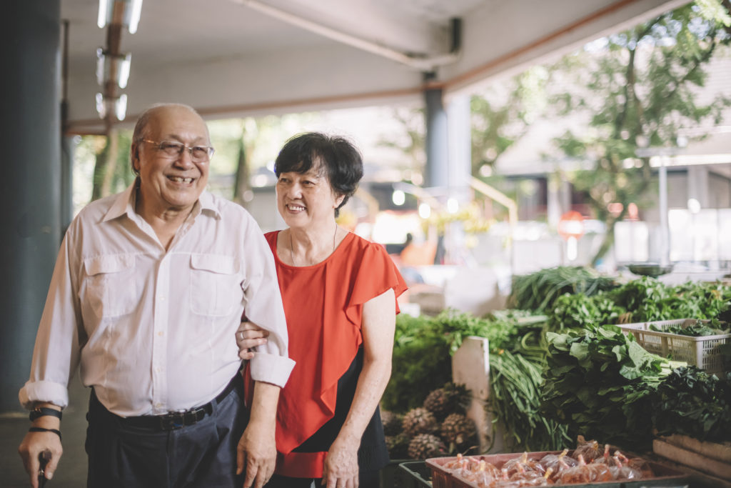 An Asian man and woman walking through an outdoor market. The man is using a cane.