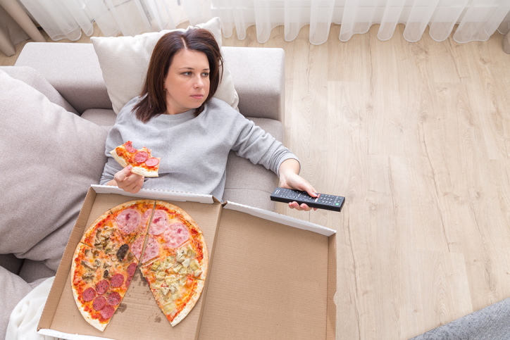 Woman laying on couch with cell phone eating pizza