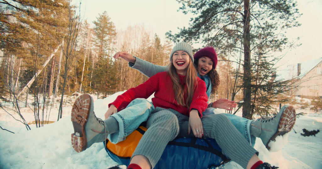 Two multiethnic excited happy beautiful friends women smile sledging down slope towards camera, winter fun slow motion. Enjoying active vacation leisure outdoors sledding down forest hill on sunny day