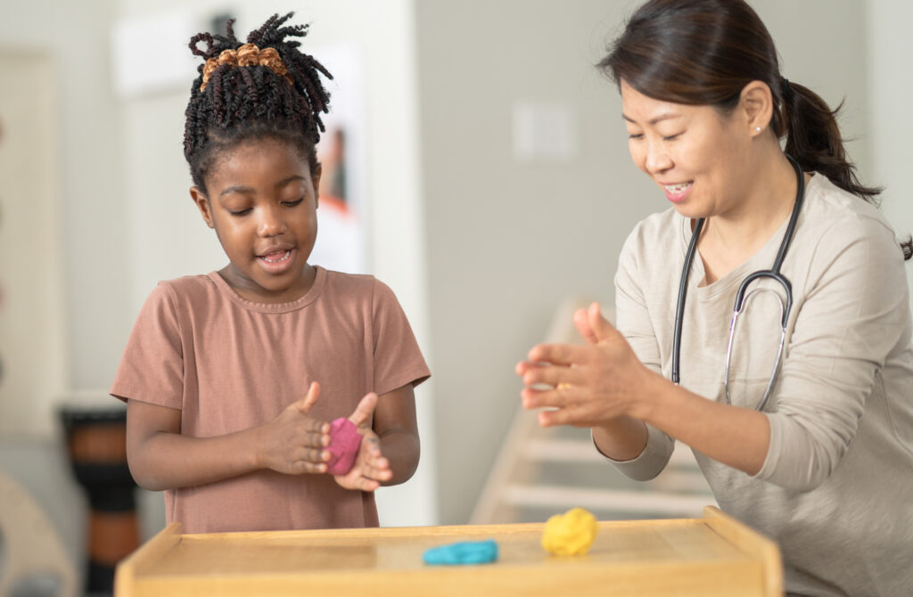 A little girl of African decent, kneels down in front of a table with her occupational therapist as they play with some colorful modeling clay. They are both dressed casually and focused on the task as they work through the therapy session together.
