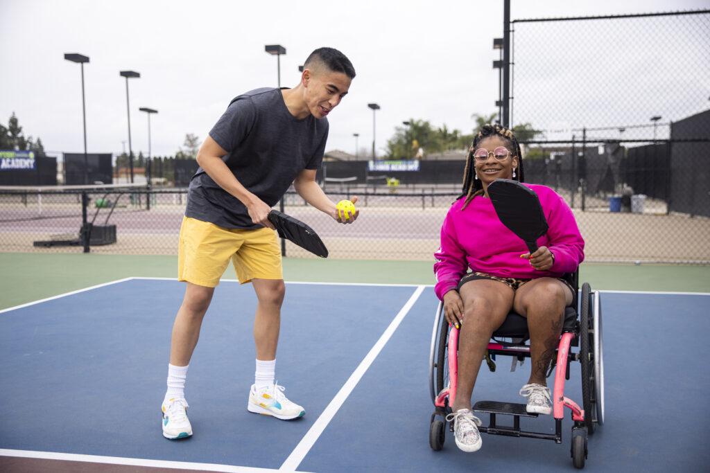 A young black disabled woman playing pickleball with her recreational rehab therapist.