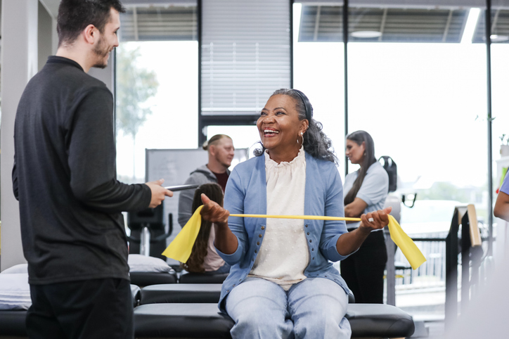 The senior woman smiles while exercising with the resistance band in the rehab clinic.